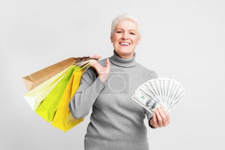 Téléchargez les photos : Femme européenne âgée gaie tenant des sacs à provisions et de l'argent ventilé, incarnant la liberté financière dans s3niorlife - en image libre de droit