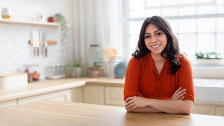 Foto de Una mujer de Oriente Medio en una blusa naranja brillante se encuentra en una cocina moderna, desprendiendo un ambiente acogedor - Imagen libre de derechos