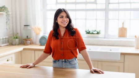 The middle eastern woman is casually leaning on a kitchen island, looking away with a smile, copy space