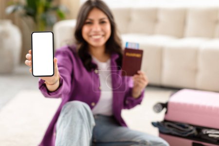 Fokussiert auf eine glückliche Reisende aus dem Mittleren Osten, die einen leeren Smartphone-Bildschirm zeigt, den Reisepass in der Hand, im Hintergrund das Wesentliche zum Reisen