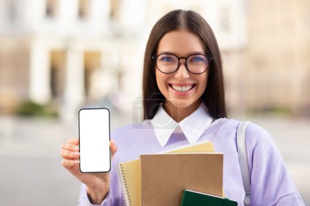 Téléchargez les photos : Joyeux jeune femme aux lunettes montrant un smartphone avec un écran vide tout en tenant des carnets, avec un fond de ville - en image libre de droit