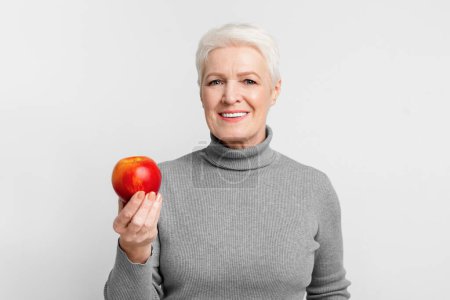 Téléchargez les photos : Femme européenne âgée souriante tenant une pomme rouge, dépeignant un mode de vie sain et s3niorlife nutrition - en image libre de droit