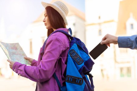 Junge Touristin, die sich auf eine Landkarte konzentriert, ist ahnungslos, als eine Diebeshand in einer urbanen Umgebung nach ihrem Handy greift