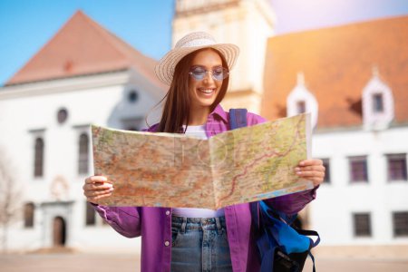 Photo for A smiling woman in a summer hat and sunglasses stands in front of a historic building, holding a large open map - Royalty Free Image