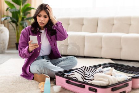 Téléchargez les photos : Inquiète femme du Moyen-Orient s'assoit sur le sol à côté de sa valise, regardant son smartphone, évoquant un sentiment d'urgence - en image libre de droit