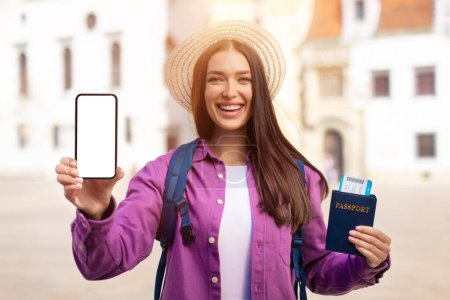 A millennial student lady poses outdoors with a passport and smartphone, ready for european travel and adventure with a smile