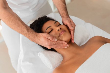 A close-up of a professional giving a relaxing neck and face massage to a black lady at a contemporary spa