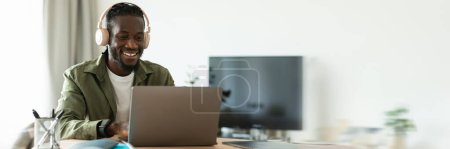 Téléchargez les photos : Heureux homme afro-américain dans un casque travaillant sur un ordinateur portable, assis au bureau à la maison et consultant des clients en ligne en utilisant PC, espace libre, bannière web - en image libre de droit