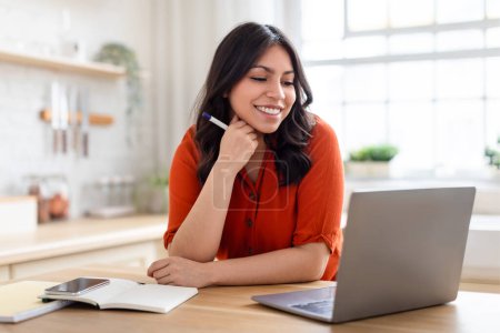 Arab young woman is seen smiling at her work, seated at a table with a laptop, notebook, and a pen in hand in a bright home setting