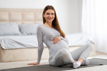 Téléchargez les photos : Une femme enceinte souriante s'assoit confortablement sur un tapis de yoga dans une chambre bien éclairée, respirant le calme et les vibrations positives - en image libre de droit