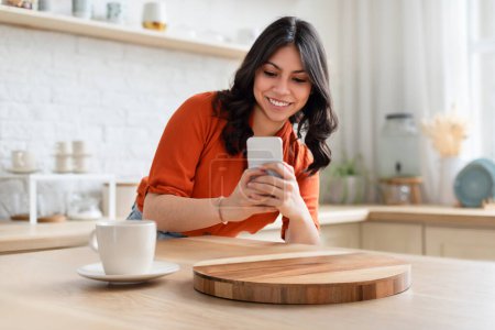 Photo for Engaged and happy middle eastern woman using her smartphone in a contemporary kitchen, possibly texting or browsing - Royalty Free Image