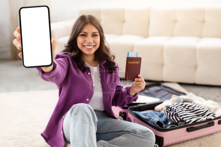 Photo for The image captures a middle eastern woman showing her smartphone and holding a travel passport beside her packed suitcase at home - Royalty Free Image