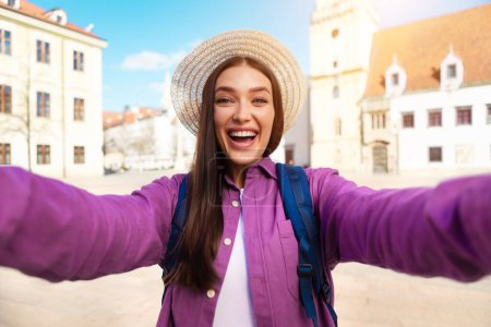 Téléchargez les photos : Jeune voyageuse souriante tient la caméra pour un selfie avec une belle place de la vieille ville et l'église en arrière-plan - en image libre de droit
