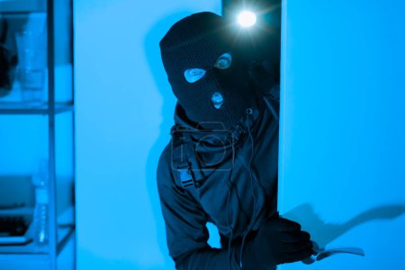 Un ladrón enmascarado con una palanca está entrando en una habitación con la intención de robar, capturado en una tenue luz azul que sugiere actividad criminal nocturna