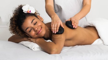 Photo for This image illustrates an african american lady experiencing blissful relaxation through a professional spa massage featuring hot stones, invoking a sense of serene indulgence - Royalty Free Image