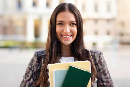 Selbstbewusste junge Studentin hält Notizbücher in der Hand und lächelt in die Kamera, Gebäude im Hintergrund, Nahaufnahme