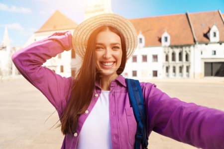 Téléchargez les photos : Une jeune étudiante prend un selfie ludique en plein air, mettant en valeur son style de vie millénaire contre l'architecture patrimoniale européenne - en image libre de droit