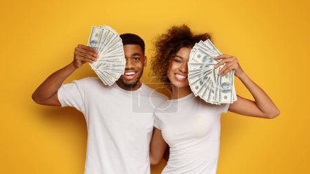 Téléchargez les photos : Un couple chanceux. Joyeux homme et femme afro-américain couvrant les visages avec des fans de dollar, fond studio orange - en image libre de droit