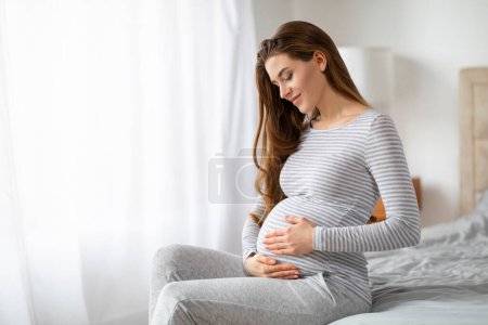Téléchargez les photos : Une dame enceinte européenne sereine en tenue confortable s'assoit de manière réfléchie, incarnant le calme et l'anticipation de la grossesse dans le confort de sa maison. - en image libre de droit