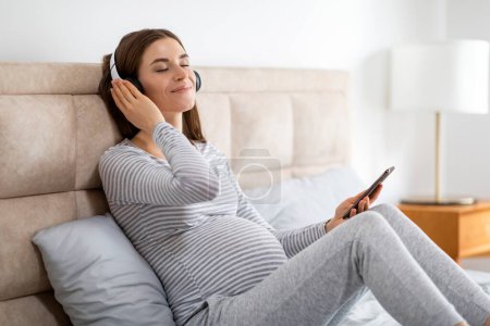 Téléchargez les photos : Une femme enceinte sereine profite de la musique avec écouteurs, assis confortablement sur un lit, tenant un smartphone - en image libre de droit