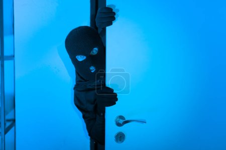Photo for A thief donning a balaclava cautiously peeks around a door, suggesting a stealthy burglar at work in an apartment late at night, capturing unlawful entry - Royalty Free Image