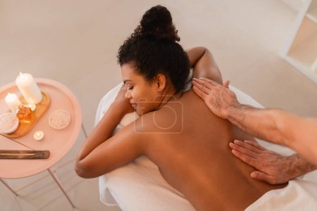 Foto de Vista de ángulo alto de la mujer afroamericana acostada en la mesa de masaje con velas alrededor disfrutando de un masaje de espalda - Imagen libre de derechos