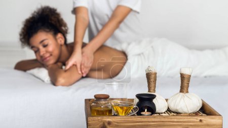 A serene African American lady enjoys a relaxing spa massage by a therapist, with essential oils and massage tools on display