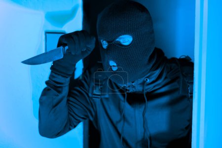 Dramatisches Bild, das einen maskierten Einbrecher zeigt, der mit einem Messer in der Hand um eine Tür lugt und den Schrecken eines Diebes verkörpert, der unter dem Deckmantel der Nacht aus einer Wohnung stiehlt