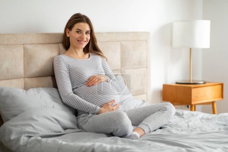 Téléchargez les photos : Une femme enceinte rayonnante en vêtements confortables sourit tout en se relaxant confortablement sur son lit, tenant son ventre - en image libre de droit