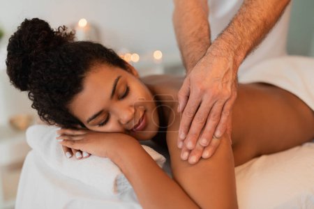 Téléchargez les photos : Une femme afro-américaine reçoit un massage doux des bras par un thérapeute, mettant en vedette l'intimité et des éléments de spa tranquilles comme des bougies - en image libre de droit