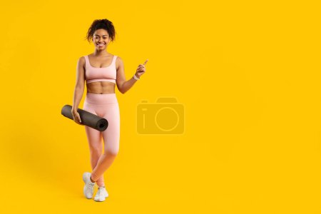 Téléchargez les photos : Une jeune femme afro-américaine en tenue de sport souriante et tenant un tapis de yoga, debout sur un fond jaune vif, espace de copie - en image libre de droit