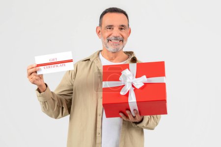 Téléchargez les photos : Un homme joyeux aux cheveux gris présente un chèque-cadeau et une boîte-cadeau rouge avec arc blanc, suggérant une occasion spéciale ou une promotion - en image libre de droit