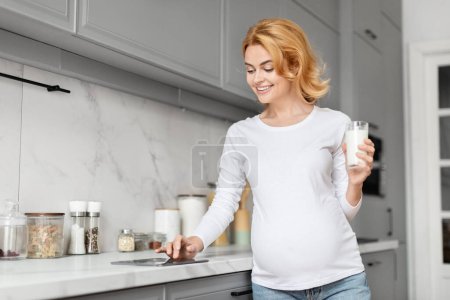 Foto de Mujer embarazada europea sonríe mientras sostiene un vaso de leche, haciendo hincapié en la importancia del calcio y la nutrición durante el embarazo en una cocina, utilizando tabletas - Imagen libre de derechos
