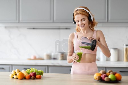 Foto de Una mujer alegre en traje de fitness está vertiendo un batido verde de una licuadora en un vaso en una cocina moderna con frutas en la mesa - Imagen libre de derechos