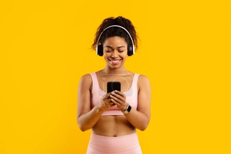 Photo for Smiling young african american woman in sporty attire with headphones using her smartphone against a bright yellow background - Royalty Free Image