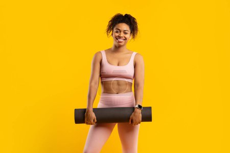 Smiling young african american woman in sportswear holding a yoga mat, set against a vibrant yellow background