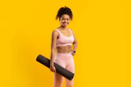 Foto de Una señora afroamericana en equipo de fitness sostiene una esterilla de yoga, aislada sobre un vibrante fondo amarillo. Sonríe cálidamente, encarnando la salud y el bienestar. - Imagen libre de derechos