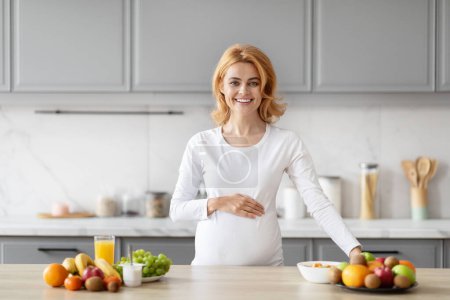 Photo for A smiling pregnant lady stands confidently in her kitchen, promoting a sense of well-being with a table of nutritious foods - Royalty Free Image