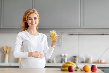 Femme enceinte européenne lumineuse boit du jus d'orange dans la cuisine, soulignant la valeur de la vitamine C pour la nutrition prénatale