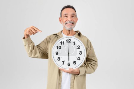 Téléchargez les photos : Un vieil homme joyeux avec une barbe tient une grande horloge murale et la pointe vers elle, debout sur un fond plat - en image libre de droit