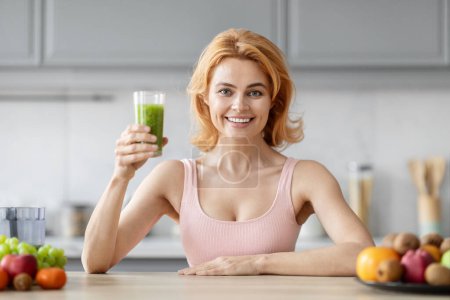 Téléchargez les photos : Une femme blonde joyeuse présentant un smoothie vert sain dans une cuisine moderne avec des fruits en arrière-plan - en image libre de droit