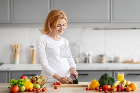 A smiling pregnant woman is slicing a cucumber in a modern kitchen, surrounded by fresh vegetables and fruits