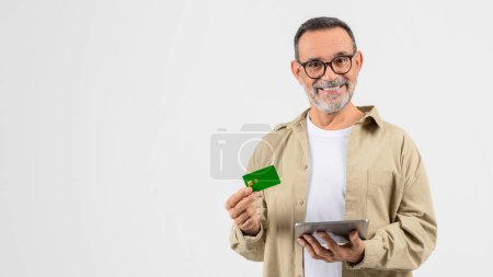 A senior man is seen isolated on white, looking at his tablet and holding a credit card, representing the elderly adapting to modern technology and online banking, copy space