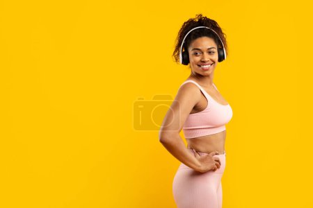 Téléchargez les photos : Fit dame noire pose en toute confiance avec les mains sur les hanches, dans les vêtements d'entraînement et écouteurs sur jaune, espace de copie - en image libre de droit