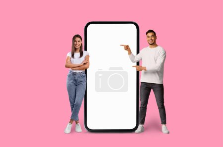 A cheerful Multiracial young couple stands next to a giant smartphone pointing and smiling at its blank screen