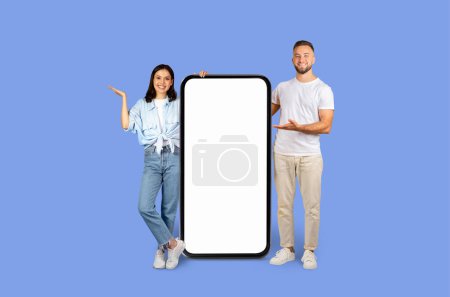 Téléchargez les photos : Un homme et une femme debout, présentant joyeusement un grand modèle de smartphone vierge à utiliser à des fins publicitaires - en image libre de droit