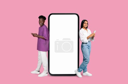 Photo for Two happy millennials Multiracial man and woman standing beside an oversized mock-up smartphone with a blank screen, ideal for advertising and apps - Royalty Free Image
