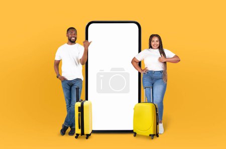 Foto de Enérgica pareja afroamericana junto con maletas enrollables que apuntan a un teléfono gigante con pantalla en blanco, perfecto para mostrar una aplicación de viaje en un fondo amarillo - Imagen libre de derechos