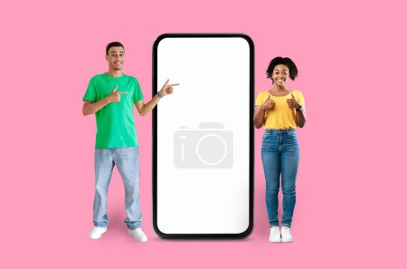 Foto de Afro-americano sonriendo hombre y mujer en ropa colorida apuntan a una enorme pantalla de teléfono inteligente en blanco con un fondo amarillo - Imagen libre de derechos