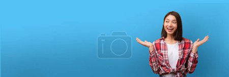 Eine junge Chinesin im karierten Hemd steht mit offenen Armen, lächelnd und aufgeregt vor blauem Hintergrund, viel Kopierraum